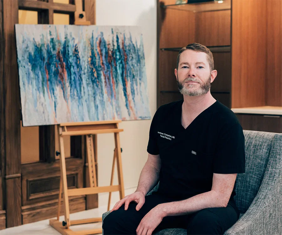 Dr. Compton sitting in a grey chair looking at the camera, resting his arms on his legs. In the background theres an easel with one of Dr. Comptons Abstract Paintings. - Andrew Compton MD in Troy, MI