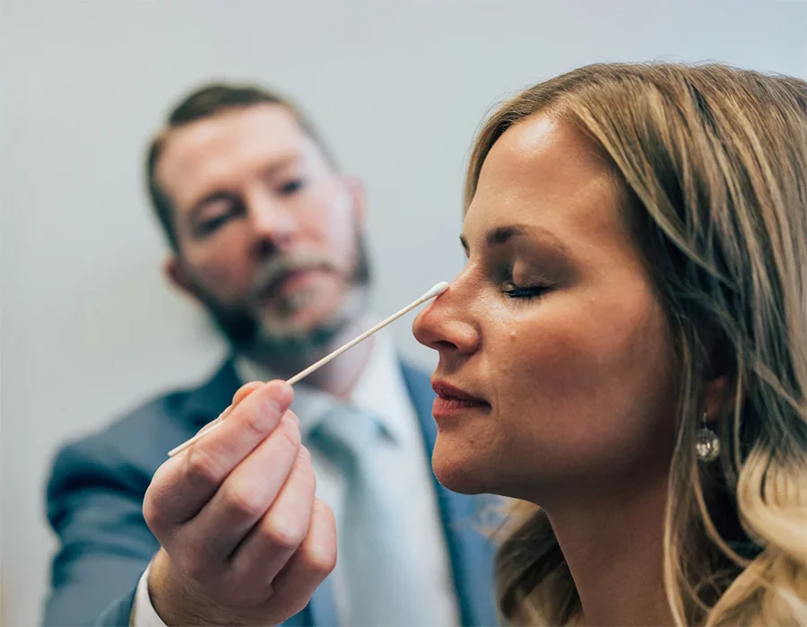 A close-up side-view shot of a female patient with her eyes closed, as a male doctor with a beard, wearing a suit and tie, gently touches her nose with a long cotton swab. This likely illustrates a consultation or examination for a nasal procedure. - Nose Procedures in Troy, MI