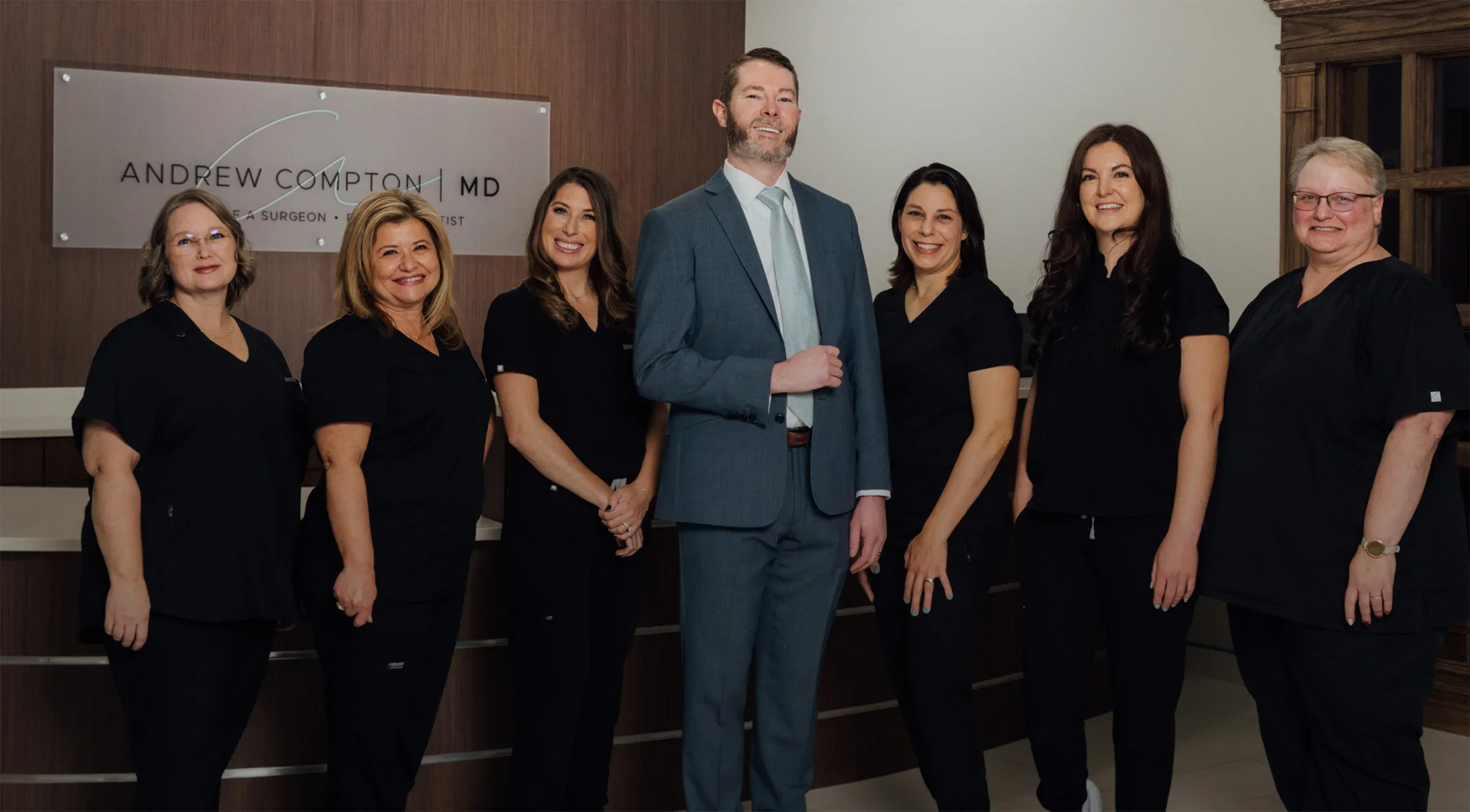 A wide shot of Dr. Compton and his team smiling at the camera in front of their reception desk. All the aestheticians are wearing black scrubs and Dr. Compton is wearing a light navy suti.