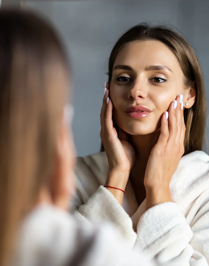 A woman wearing a light beige robe looks at her reflection in a mirror, gently touching her face and neck with a pleasant, focused expression. Her skin appears smooth, and a thin red bracelet is visible on her wrist. A blurred figure, likely her own, is seen in the reflection against a muted gray background. - Buccal Fat Removal in Troy, MI