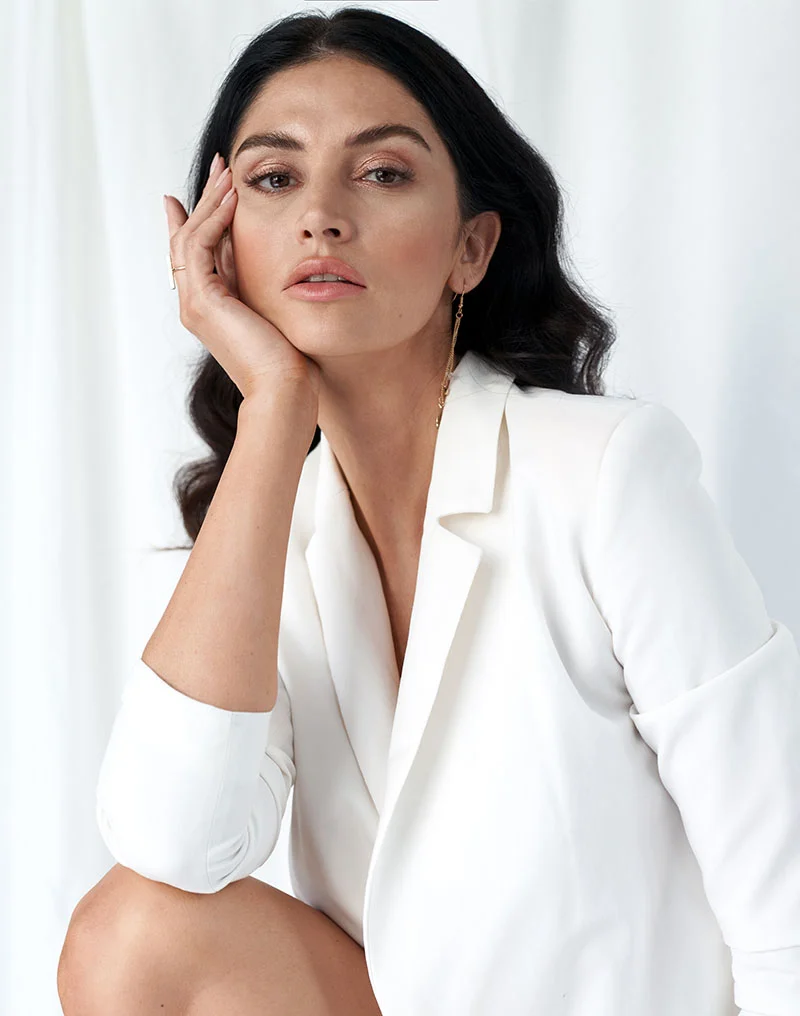 A beautiful woman with long, dark, wavy hair and makeup, wearing a white blazer, poses looking directly at the camera with a serious expression. She is resting her chin on the back of her hand. She is wearing a delicate gold drop earring. The background is a bright white studio setting with soft lighting. - Chin Augmentation in Troy, MI