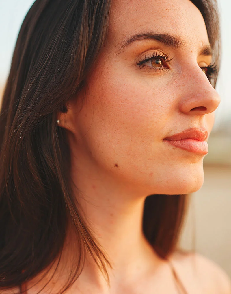 A close-up profile portrait of a beautiful young woman with long dark hair, freckled skin, and defined eyebrows. She is looking off to the side, and the warm, soft outdoor lighting highlights her features. She is wearing a small stud earring and a tank top. - Chin Neck Liposuction in Troy, MI
