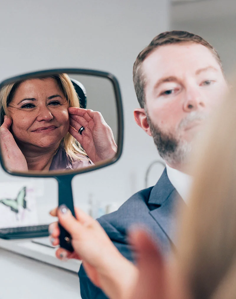 A close-up shot showing a woman with blonde hair looking happily at her reflection in a handheld mirror. The reflection shows her touching her cheek and smiling. Dr. Compton with a beard, wearing a suit, is blurred in the foreground, looking over her shoulder into the mirror, likely reviewing the results of a consultation or procedure. - Deep Plane Facelift in Troy, MI