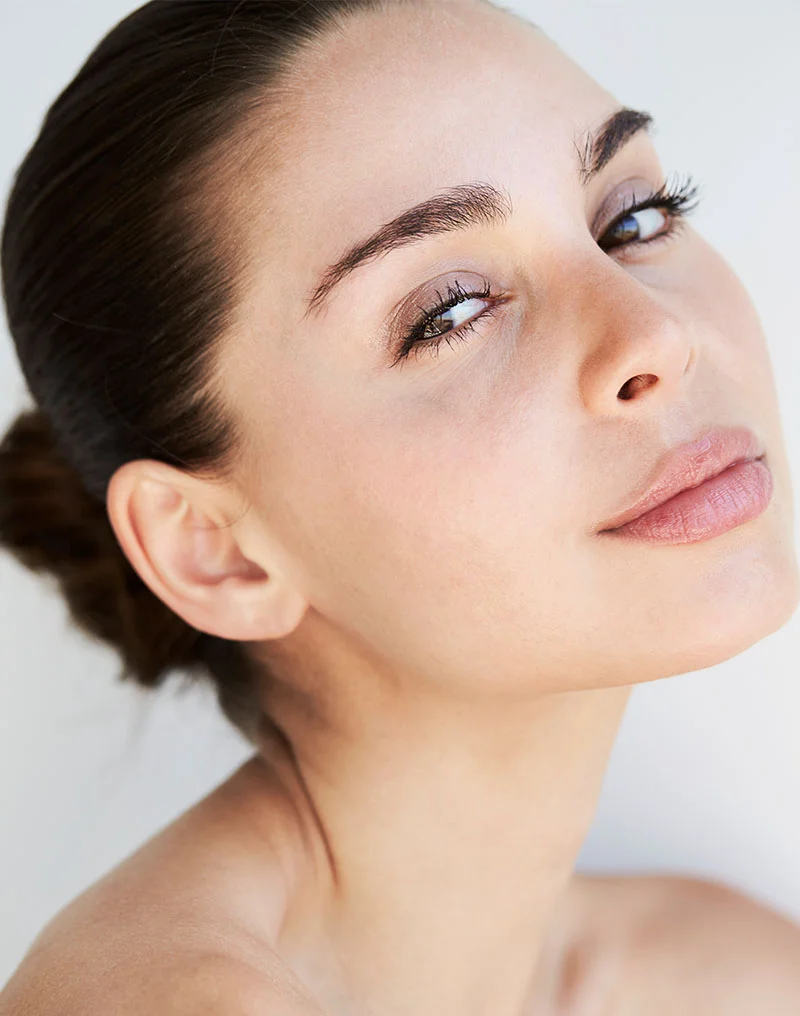 A close-up, high-angle portrait of a beautiful woman with her dark hair pulled back in a bun. She has full lips and defined eyebrows, and she is looking up and to the side with a sultry expression. Her skin is radiant, and her shoulders are bare, suggesting an aesthetic or spa setting. The background is bright white. - Dermal Fillers in Troy, MI