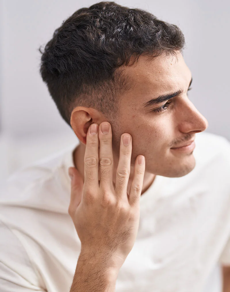 A close-up profile shot of a young man with dark, wavy hair and a slight smile, wearing a white t-shirt. He is gently touching the side of his face and his ear with his right hand, looking off to the right. The background is a bright, solid light color. - Earlobe Reduction in Troy, MI