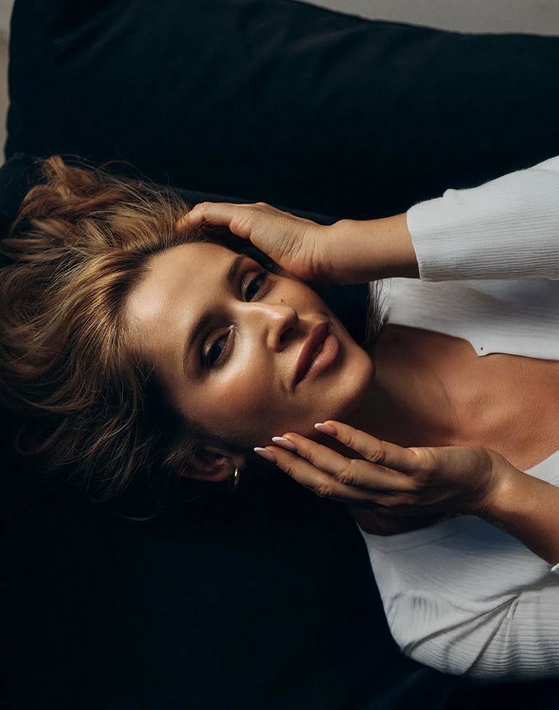 A dramatic, close-up portrait of a woman with blonde hair, lying on a dark surface and wearing a white top. She is looking up at the camera and gently touching the sides of her face with both hands, highlighting her facial contours. The lighting creates a contrast between her glowing skin and the dark background. - Facial Implants in Troy, MI