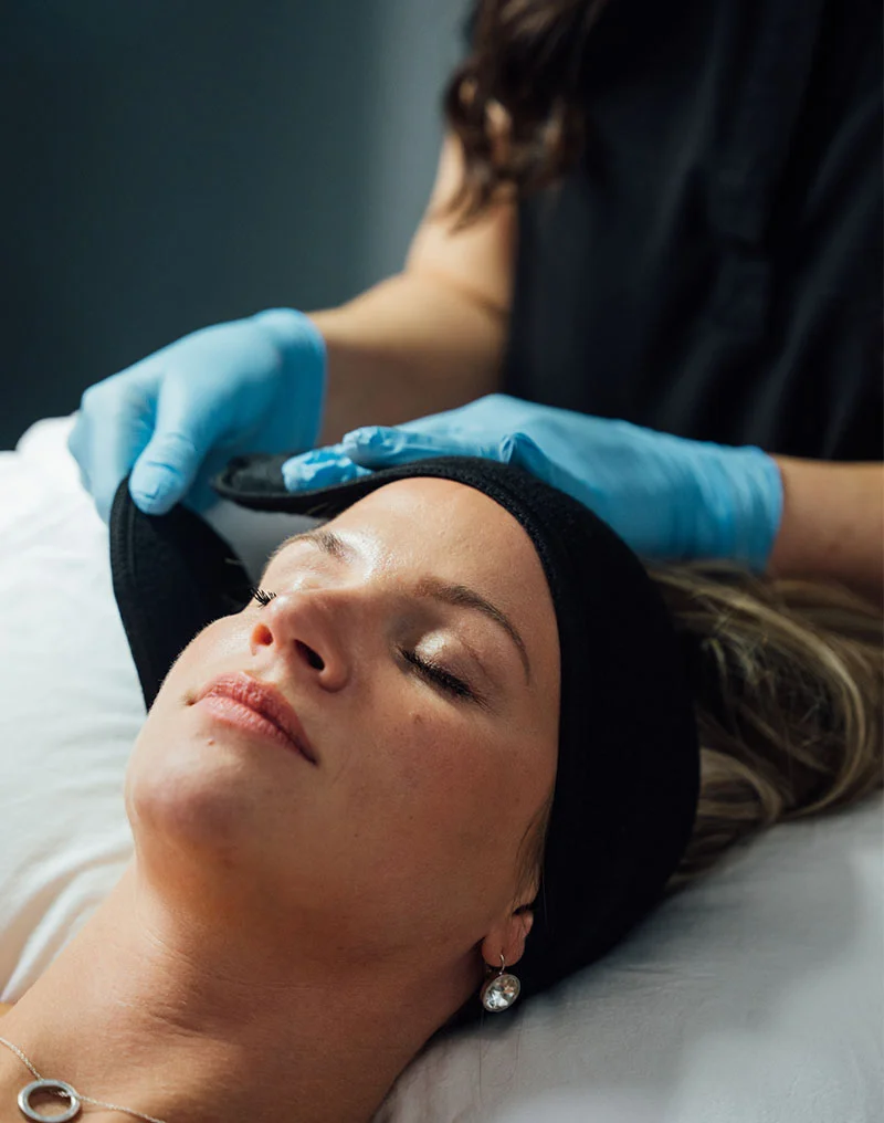 A close-up of a female patient lying down with her eyes closed, wearing a black headband for a facial treatment. An aesthetician, wearing blue gloves, is gently adjusting the headband on the patient's forehead. The patient has blonde hair and is wearing a silver necklace and a dangling silver earring. - Fractional Plasma in Troy, MI