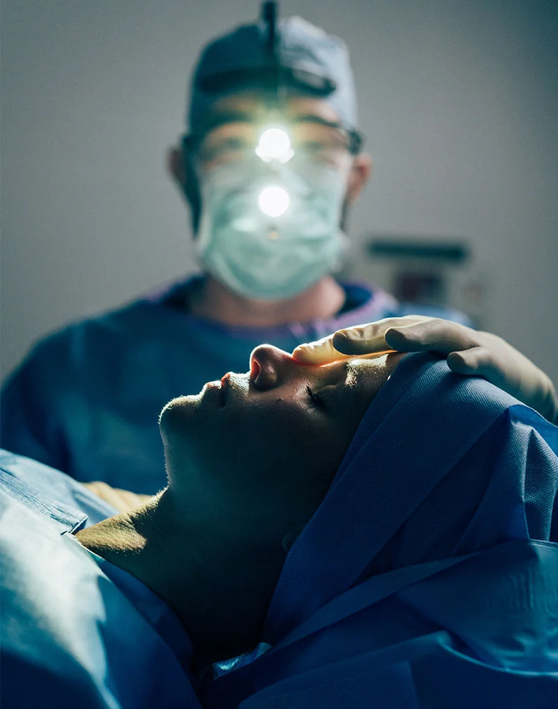 A dimly lit photo shows a patient, draped in blue surgical cloth, lying on an operating table, with their face illuminated by the bright headlamp of the surgeon standing behind them. Dr. Compton, the surgeon, is out of focus and wearing a surgical mask, cap, and headlamp, while his gloved hand rests gently on the patient's forehead, likely during a functional rhinoplasty procedure. - Functional Rhinoplasty in Troy, MI