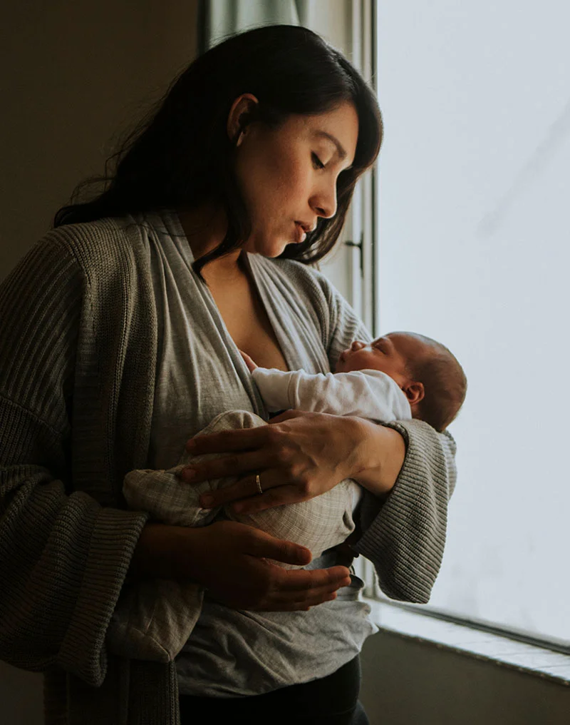 A soft-focus, intimate portrait of a mother with long dark hair, wearing a gray ribbed cardigan, holding her newborn baby close to her chest. The baby is wearing a white and light-colored outfit and appears to be sleeping. They are standing near a window with bright natural light coming in from the side. - Infant Earlobe Molding in Troy, MI