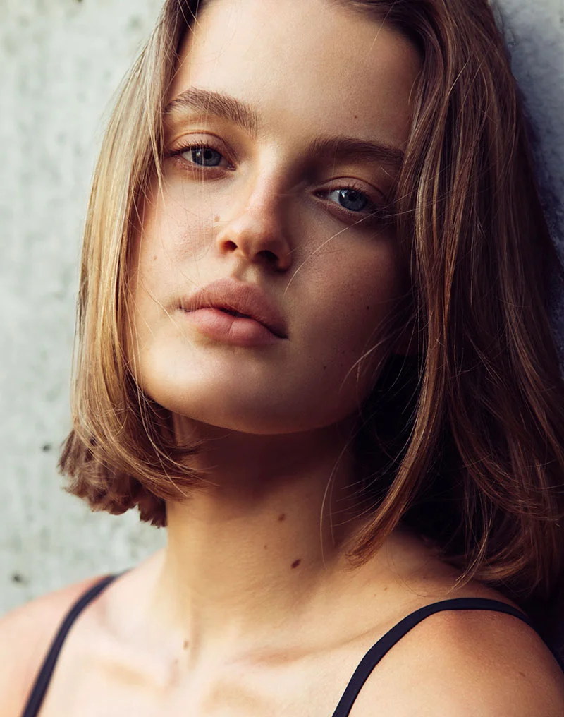 A close-up vertical portrait of a young woman with a straight, shoulder-length bob hairstyle and light eyes, leaning against a rough, gray concrete wall. She is looking directly at the camera with a neutral expression and slightly parted lips. She is wearing a dark, thin-strapped top. The lighting highlights her features and gives a slightly moody feel to the image. - Lip Fillers in Troy, MI