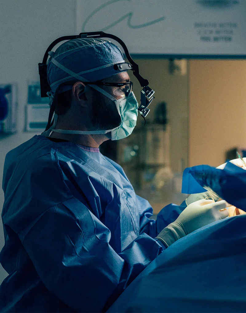 Dr. Compton, a male surgeon, is shown in profile in an operating room, leaning over a patient. He is wearing blue surgical scrubs, a surgical cap, a face mask, glasses, and a surgical loupe with a headlamp. His gloved hands are visible working on the patient, who is draped in blue surgical cloth. The lighting is focused on the surgical field. - MOHs Reconstruction in Troy, MI