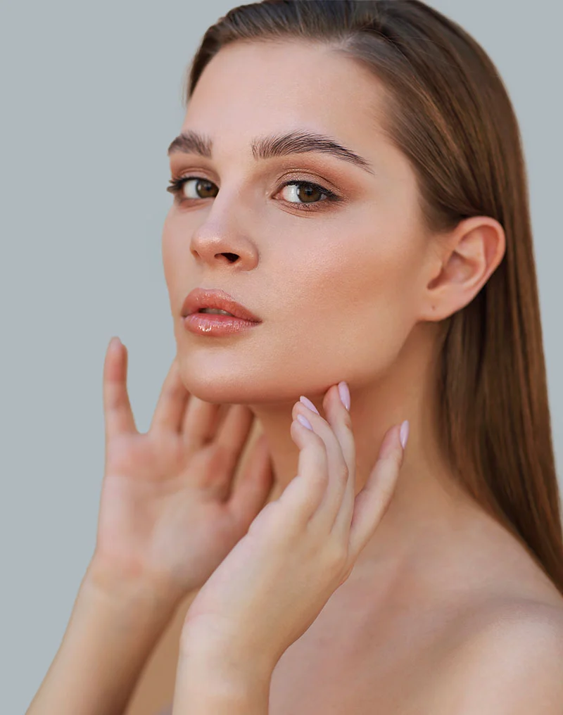A head and shoulder studio portrait of a woman with long, straight brown hair, looking directly at the camera. Her face is soft and clear, with subtle makeup highlighting her eyes and lips. She is touching the sides of her chin and jawline with both hands, palms facing inward, drawing attention to her neck and lower face. The background is a plain, light grayish-blue. The lighting is soft and even. - Morpheus8 in Troy, MI
