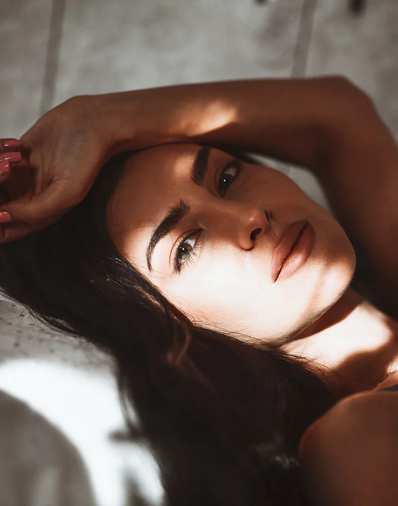 A dramatic, close-up photo of a young woman lying down, looking directly at the camera with an intense gaze. She has dark hair and one arm is draped over her forehead. Her face is highlighted by a strong ray of sunlight and shadow, emphasizing her eyes and lips. - Nano Fat Stem Cells in Troy, MI