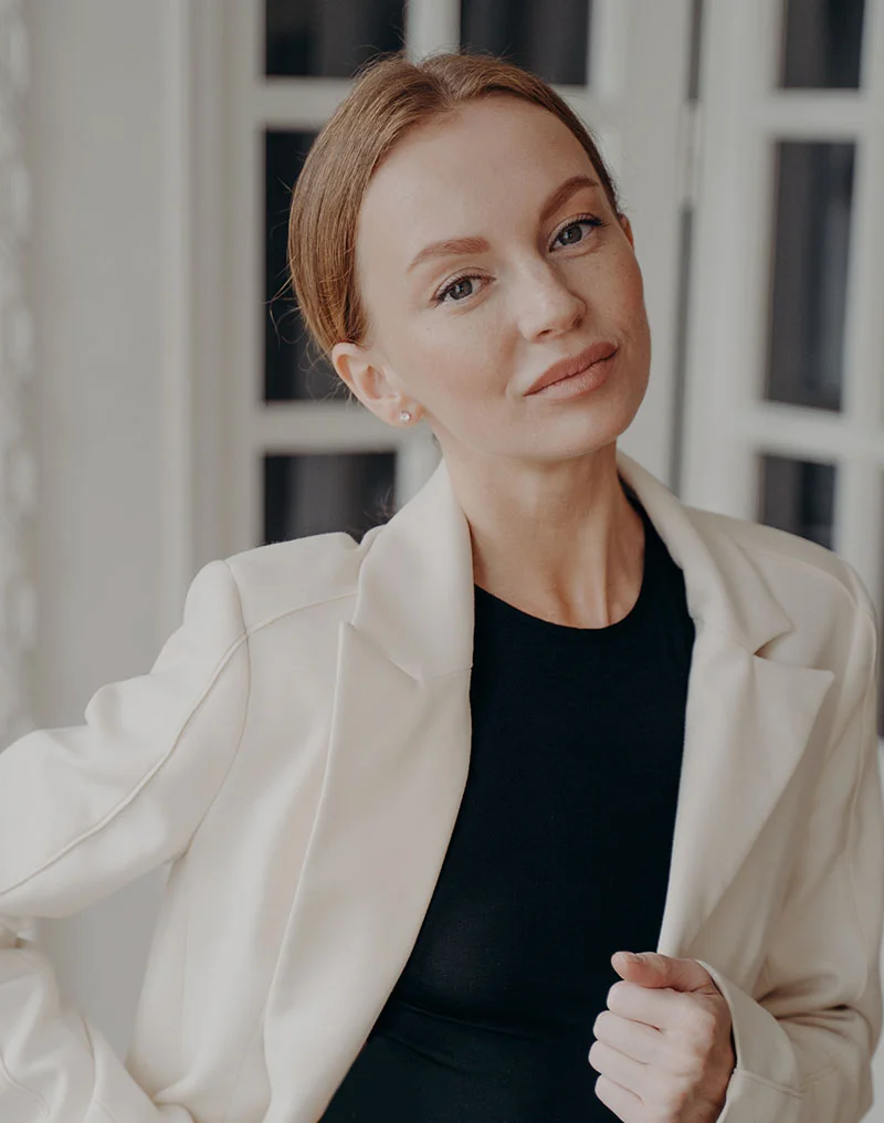 A portrait of a beautiful woman with light reddish-blonde hair pulled back, wearing a cream or white blazer over a black top. She is looking directly at the camera with a calm expression. The background is an interior space with bright white french doors or windows. - Neck Lift in Troy, MI