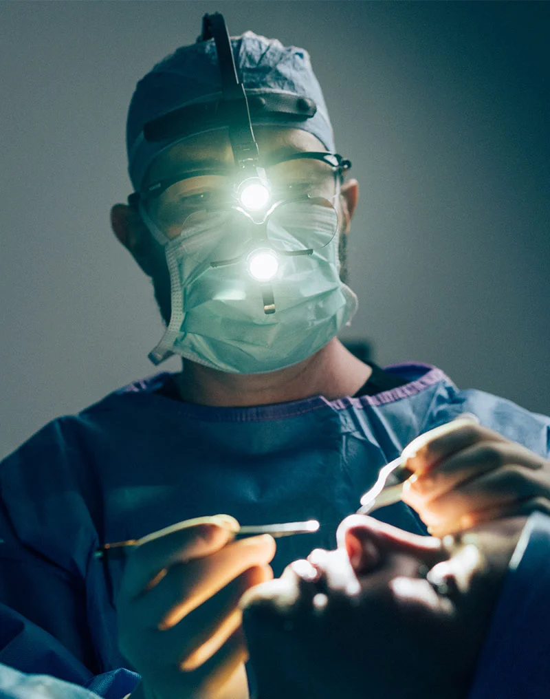 A dramatic, low-angle close-up of Dr. Compton, a male surgeon, performing a procedure. He is wearing blue surgical scrubs, a surgical cap, a mask, glasses, and a surgical loupe with a very bright headlamp that illuminates the area of the patient's nose. His gloved hands are holding surgical instruments above the patient's face, which is partially visible in the foreground. - Revision Rhinoplasty in Troy, MI